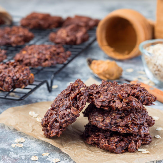 A pile of dark chocolate oat cookies resting on a rustic wooden table with scattered oats, a metal cooling rack, and a terracotta cup in the background.